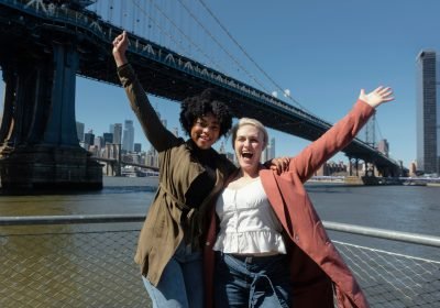 Imagem mostrando duas mulheres felizes e sorridentes, próximas a uma ponte famosa nos EUA, após conseguirem seu green card.